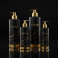 Four matte black TITAN shower gel bottles with gold pumps, bases, and gold swirl accents on a wet reflective surface.